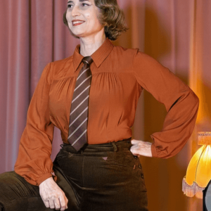 Frau mit kupfer farbener Bluse im Stil der 30er Jahre und Krawatte. Im Hintergrund pink Samtvorhang
