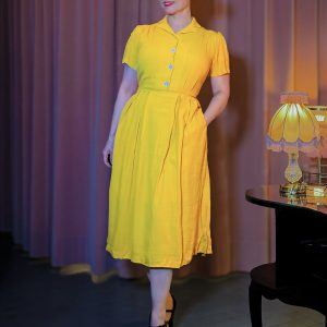 Senfgelbes Hemdblusenkleid im Stil der 40er Jahre mit kontrastierenden hellblauen Knöpfen und weitem Rockteil, elegant kombiniert mit schwarzen T-Strap-Schuhen vor einem stimmungsvollen Bühnenambiente – Hazel's Boudoir