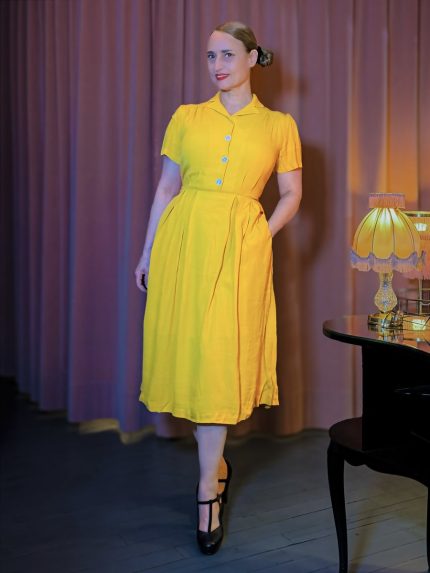 Senfgelbes Hemdblusenkleid im Stil der 40er Jahre mit kontrastierenden hellblauen Knöpfen und weitem Rockteil, elegant kombiniert mit schwarzen T-Strap-Schuhen vor einem stimmungsvollen Bühnenambiente – Hazel's Boudoir